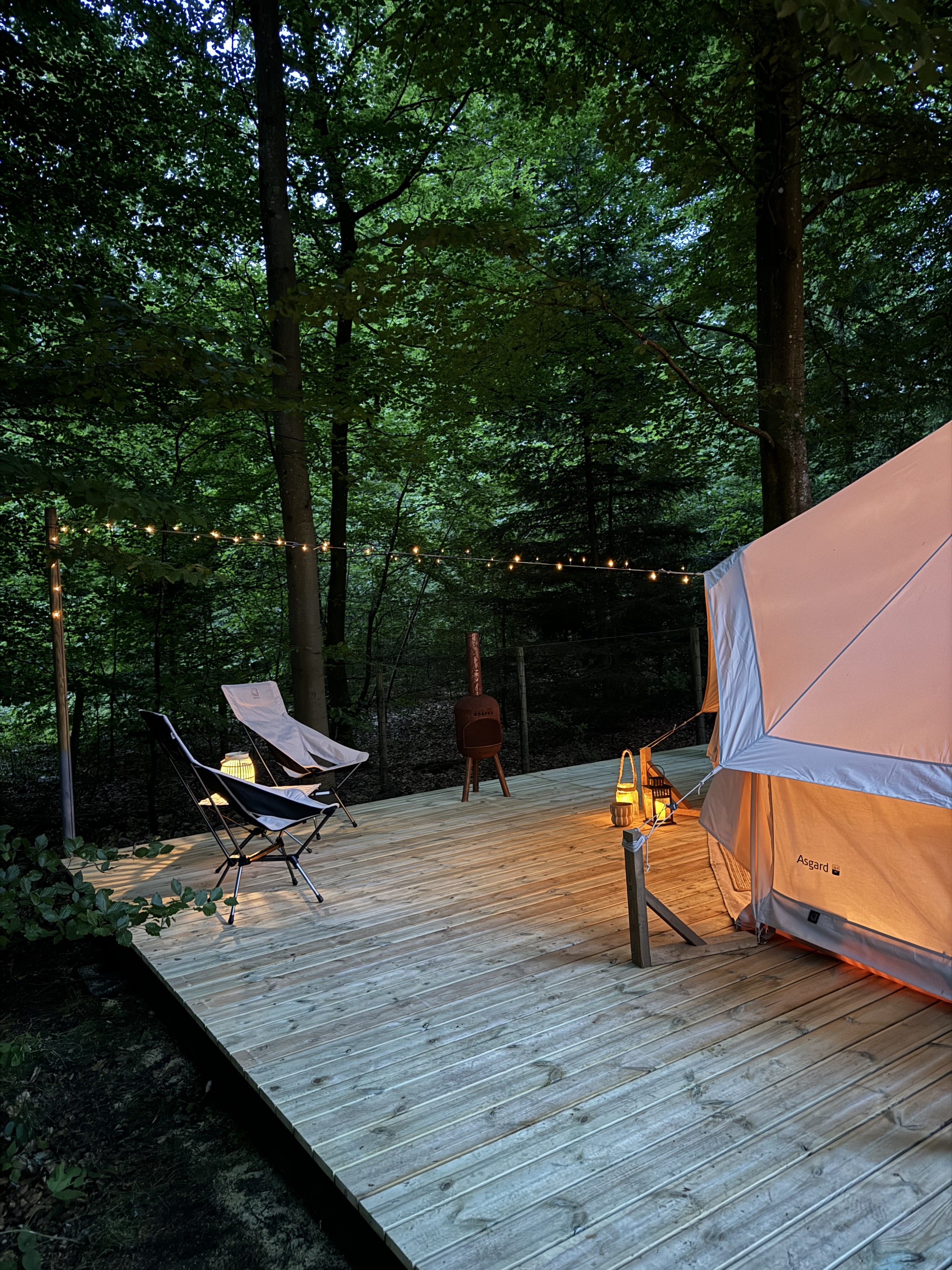 Glampingtelt på træterrasse foran skov i aftenlys, oplyst af lyskæder og lanterner der skaber en hyggelig stemning